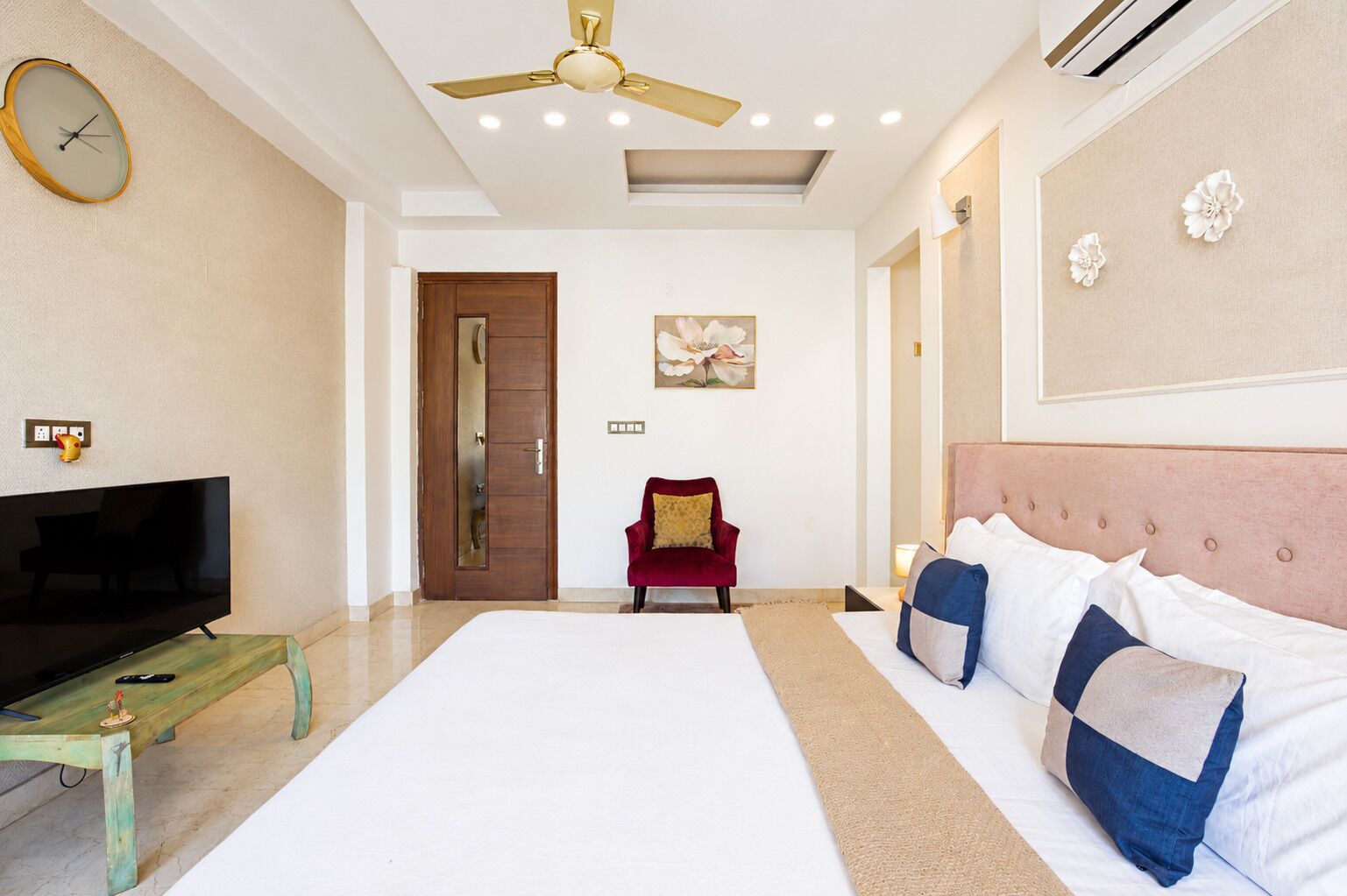 A hotel room with a bed, a TV, a red chair, a ceiling fan, and a painting on the wall.