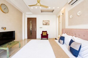A hotel room with a bed, a TV, a red chair, a ceiling fan, and a painting on the wall. - Blossom by Redolive 3bhk Golf Course Ext (Gurugram)