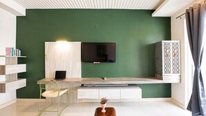 Ein modernes Wohnzimmer mit grüner Wand, einem aufgehängten Fernseher, einem Holzkonsolentisch, einem grünen Sessel, einem kleinen Tisch mit einer Vase und einem Bücherregal.