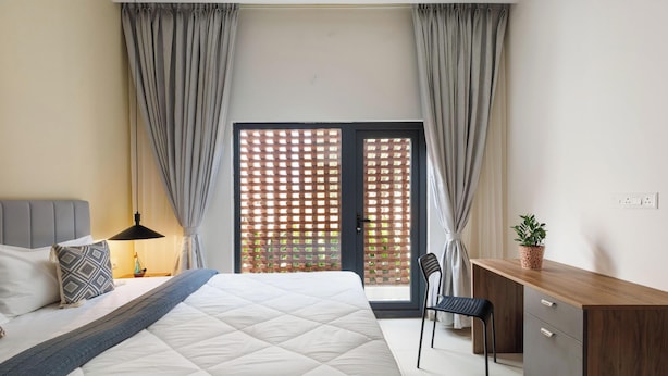 A bedroom with a bed, bedside table, lamp, desk, chair, and a view through a window with curtains. - Aero Rosewood by Red Olive 3bhk 5min Airport (Bengaluru)