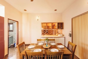 A dining area with a wooden table set for six, surrounded by chairs, with a kitchen in the background. - Bluemirage Villapoolgardenjacuzzi30min Airport (Doddaballapur)