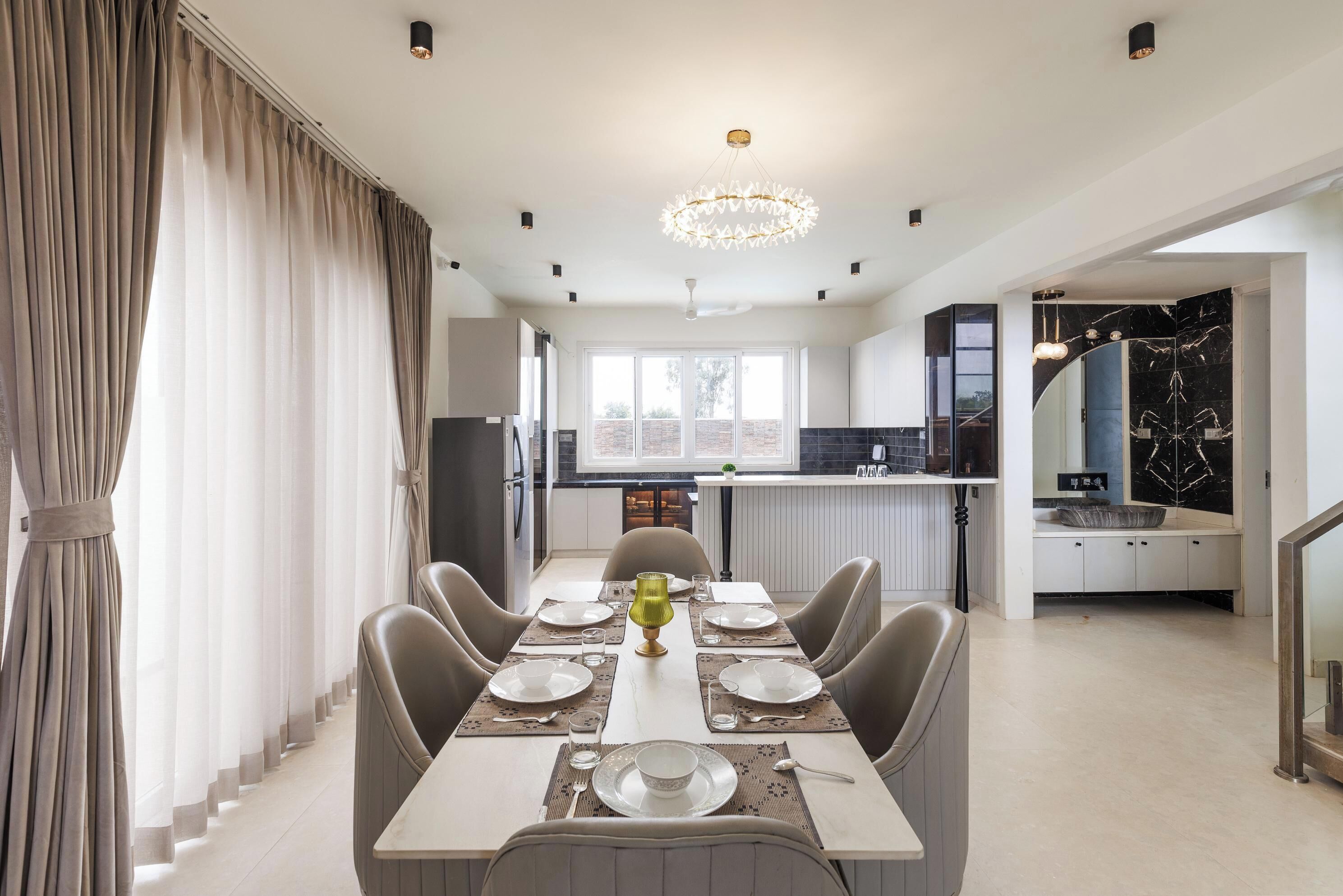 Un comedor moderno con una larga mesa dispuesta para seis personas, una araña y vistas a la cocina y la sala de estar.