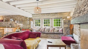 A cozy living room with a stone wall, a wooden ceiling, a red sofa, a wooden table, and a window with a view of greenery. - Stone house for 5, garden, beach 700m away (Sarzeau)