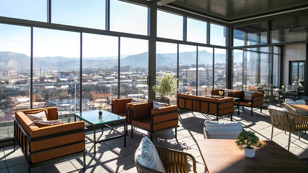 A spacious rooftop terrace with large windows offering a cityscape view, furnished with sofas, tables, and chairs.