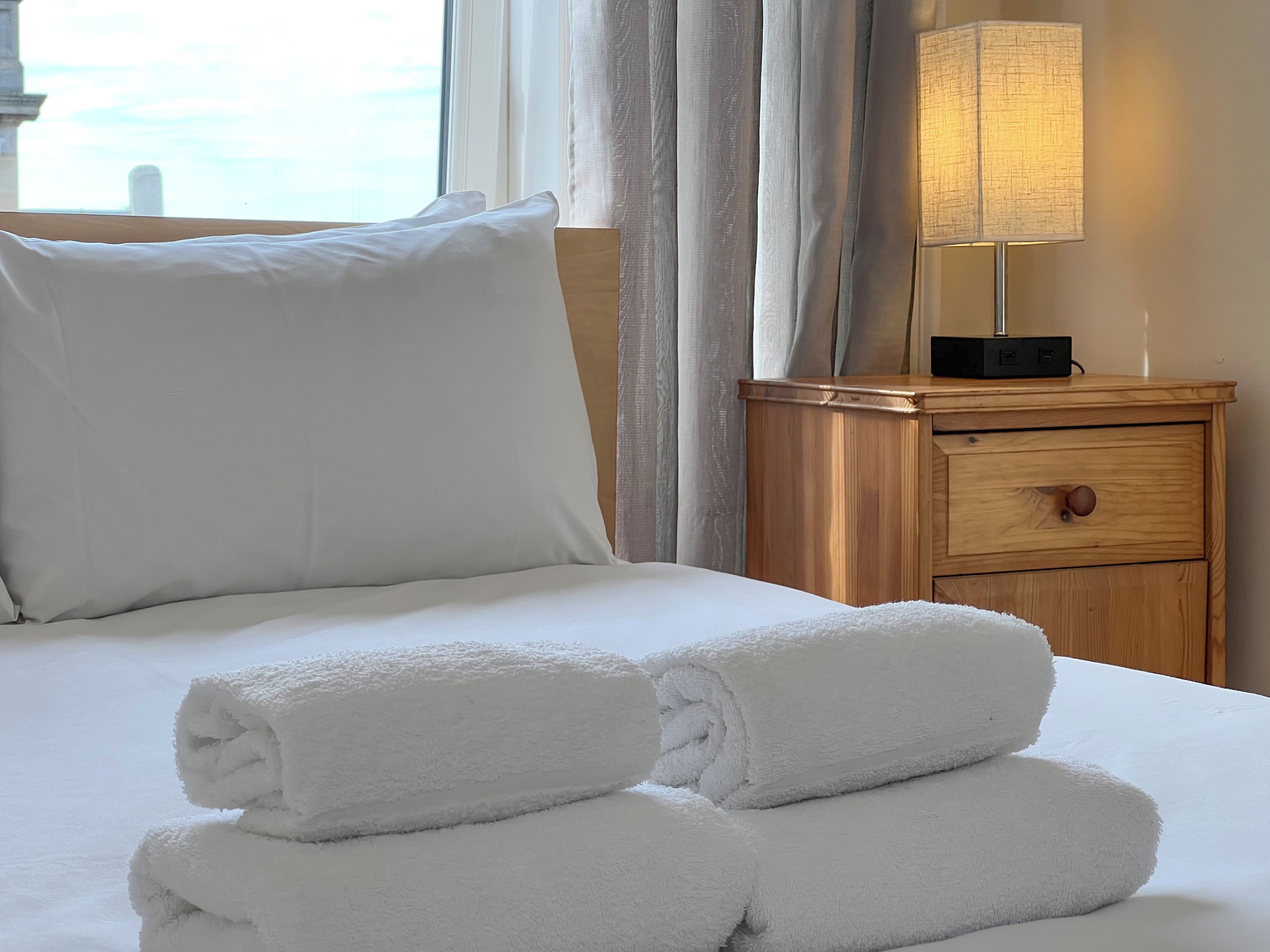 A neatly made bed with white linens and rolled towels, a bedside lamp, and a wooden nightstand with a drawer.