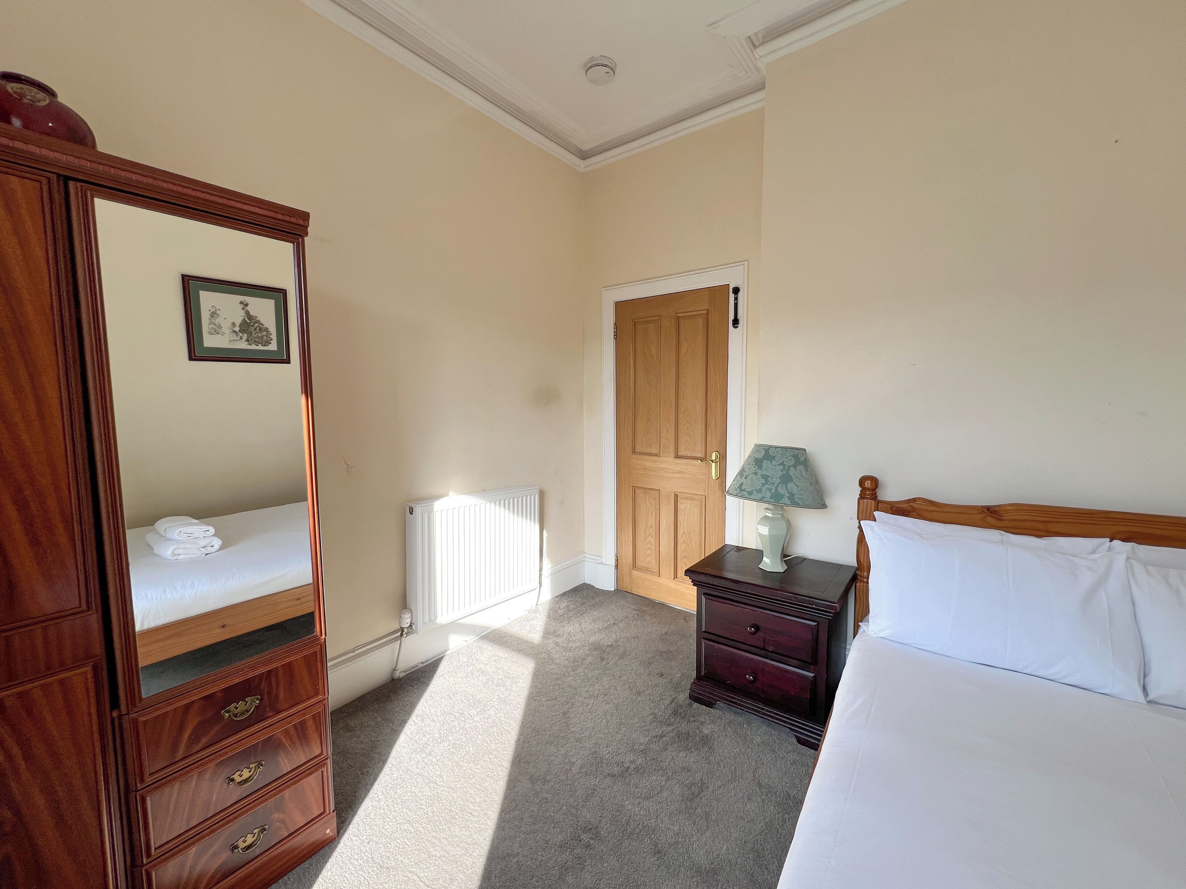 A bedroom with a wooden dresser, a bedside table with a lamp, a radiator, and a door.