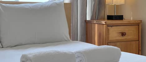 A neatly made bed with white linens and rolled towels, a bedside lamp, and a wooden nightstand with a drawer.
