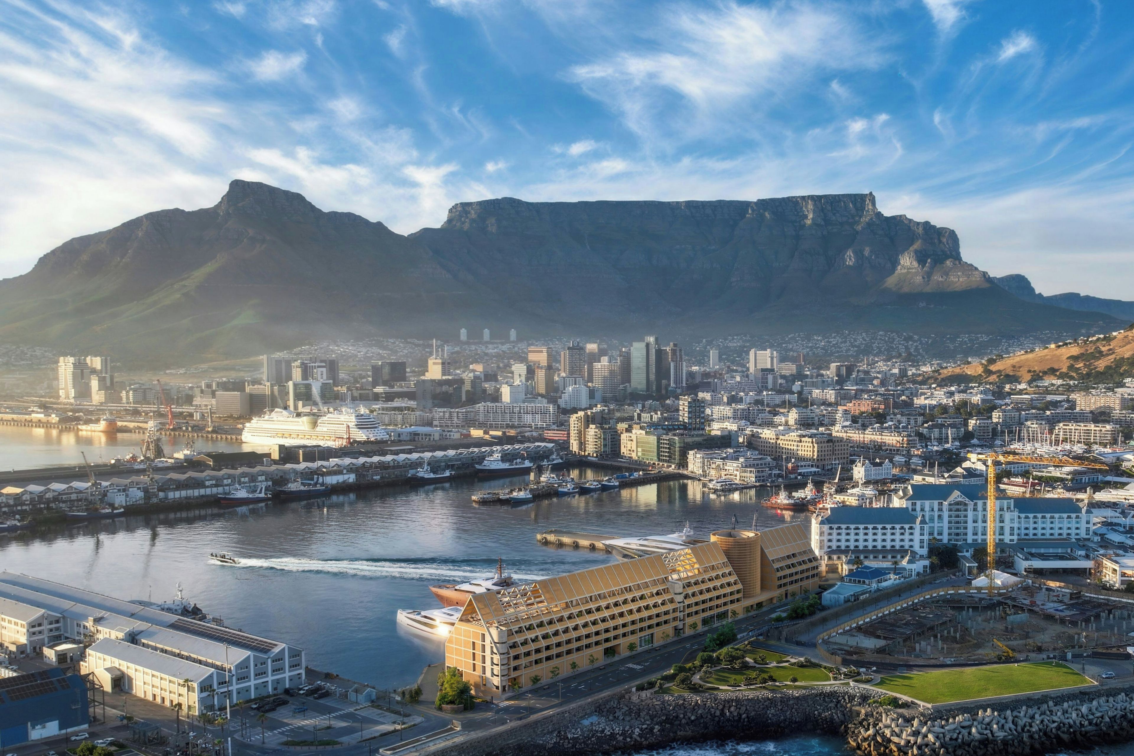Un paysage urbain côtier avec une montagne imposante en arrière-plan, un port de plaisance avec des bateaux et des bâtiments modernes.