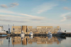 Una marina con varios yates amarrados, un edificio moderno con fachada de diseño geométrico y una superficie acuática tranquila que refleja las estructuras.