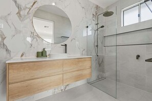 Een moderne badkamer met een marmeren wand, een ronde spiegel, een houten wastafelmeubel, een douchecabine van glas en een raam.