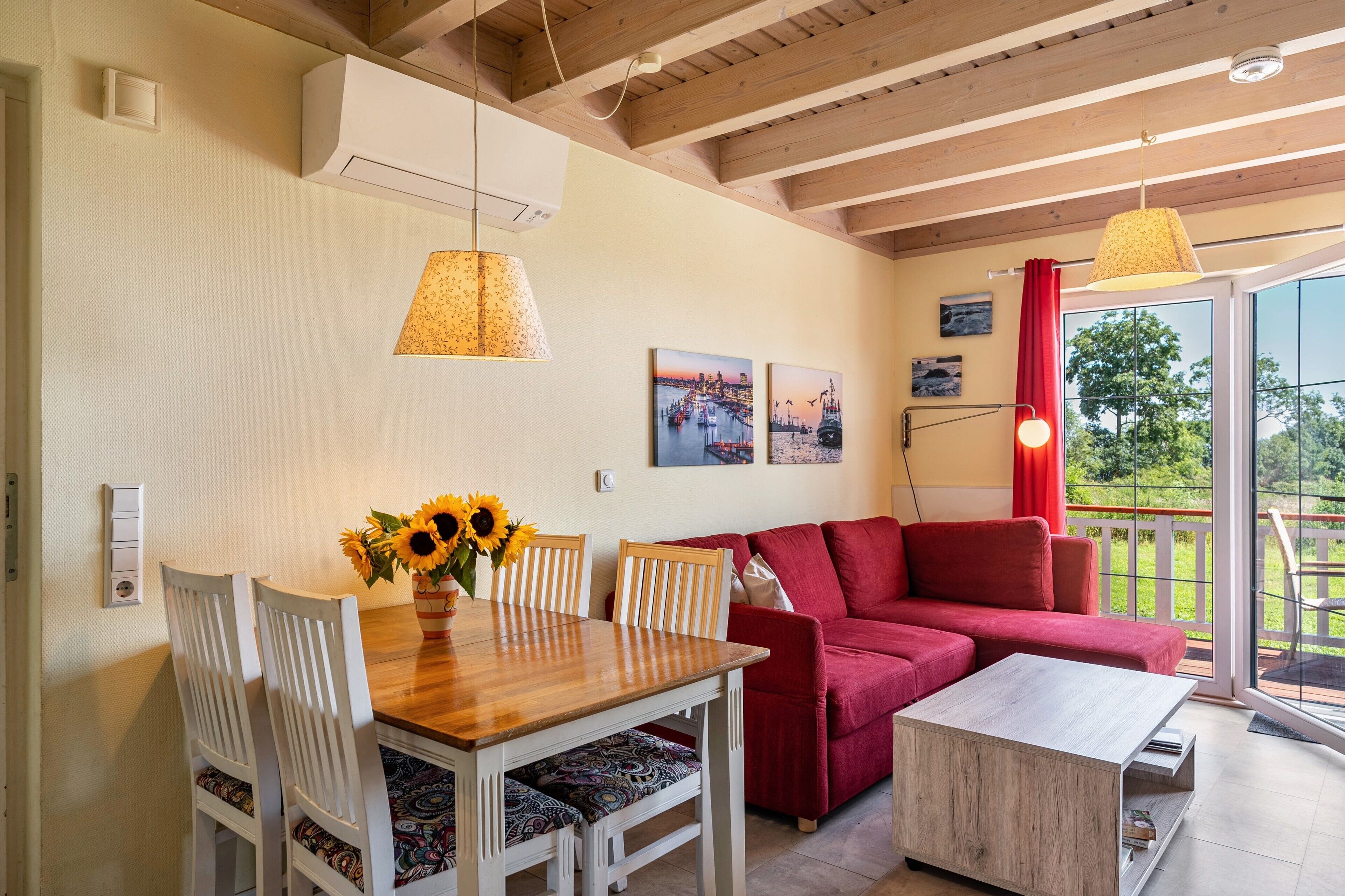 Un coin repas avec une table en bois, des chaises blanches et un vase rempli de tournesols. Un canapé rouge et une petite table en bois sont placés contre le mur.