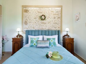 A bedroom with a stone-patterned headboard, a bed with a blue quilt, two bedside tables with lamps, and a small decorative wreath. - 3-bed Armeni Mountain Oasis (Rethymno)