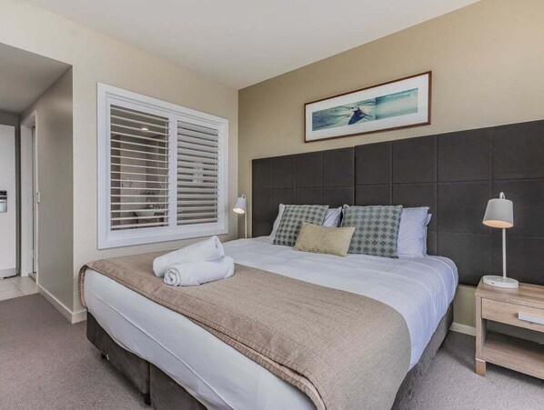 A bedroom with a bed, a nightstand, a lamp, a window with blinds, and a framed picture on the wall. - The Pool Resort Kingscliff - Two Bedroom (5307/5308) with garden view - Holiday Management Co. (Tweed)
