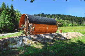 A wooden cylindrical structure with a roof, situated on a grassy area with trees in the background. - Glamping 'Sleeping Barrel' in the Black Forest (Alpirsbach)