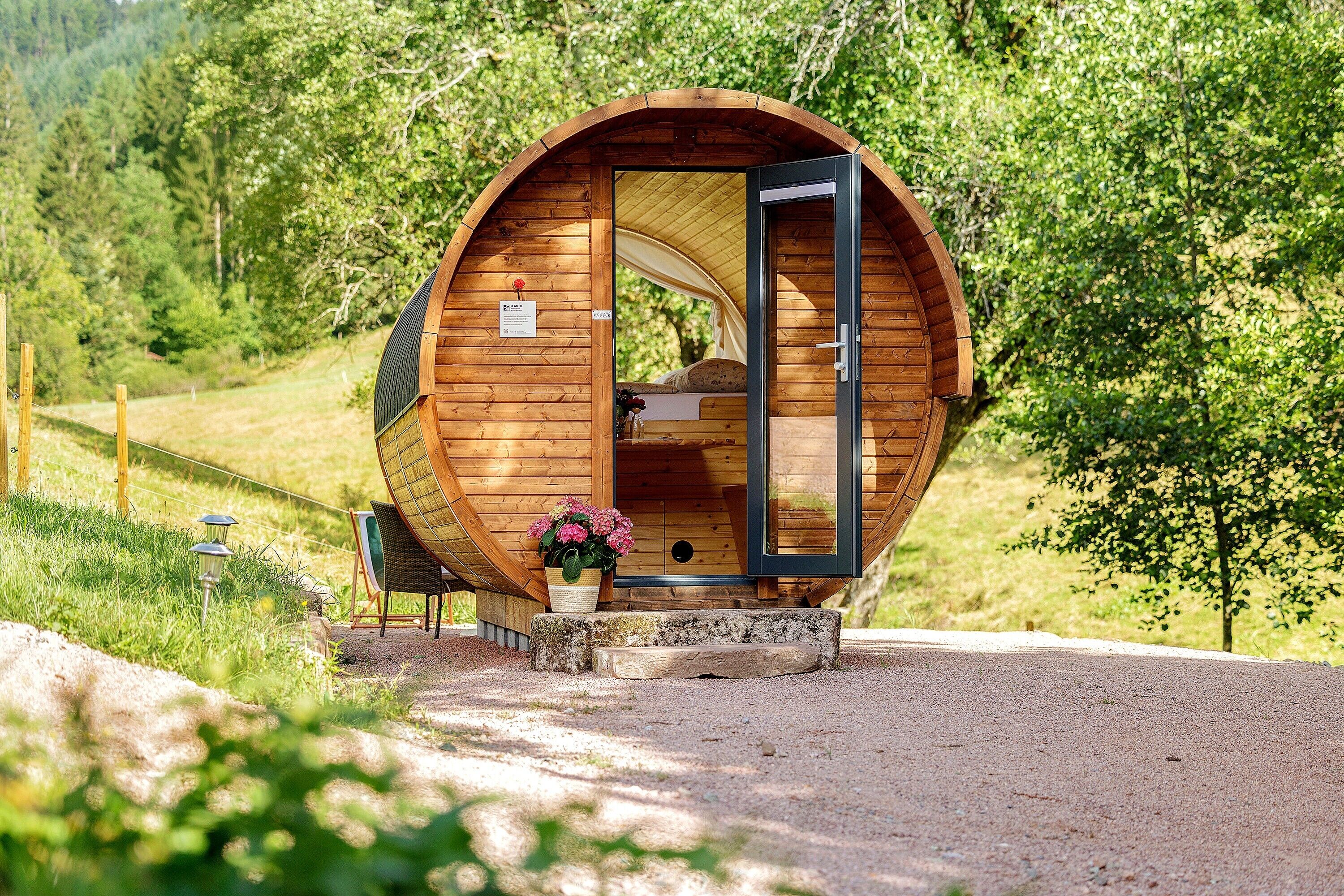 Una estructura circular de madera con puerta abierta, una cama en su interior y un pequeño porche con una maceta.