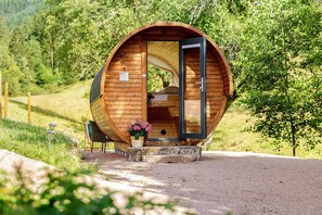 A wooden, circular structure with an open door, a bed inside, and a small porch area with a flower pot. - Glamping 'Sleeping Barrel' in the Black Forest (Alpirsbach)