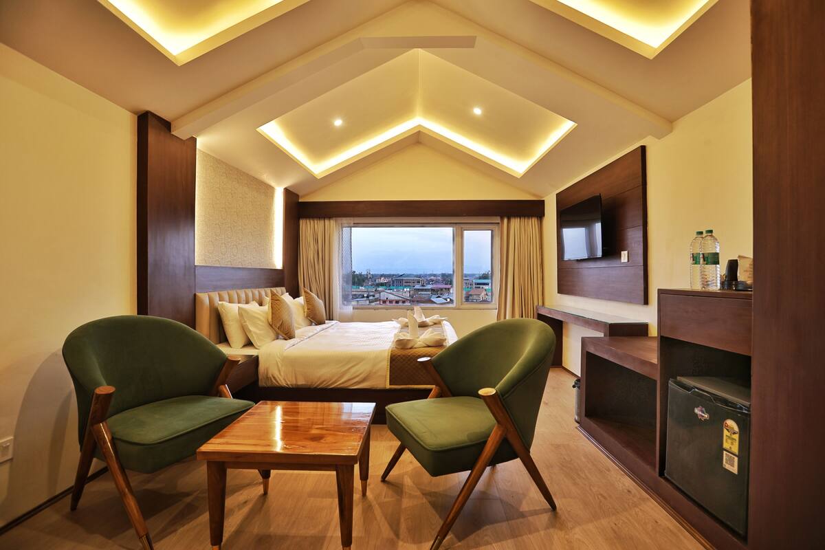 Quarto de hotel com uma cama, duas cadeiras verdes, uma mesa de madeira, uma televisão e um refrigerador.