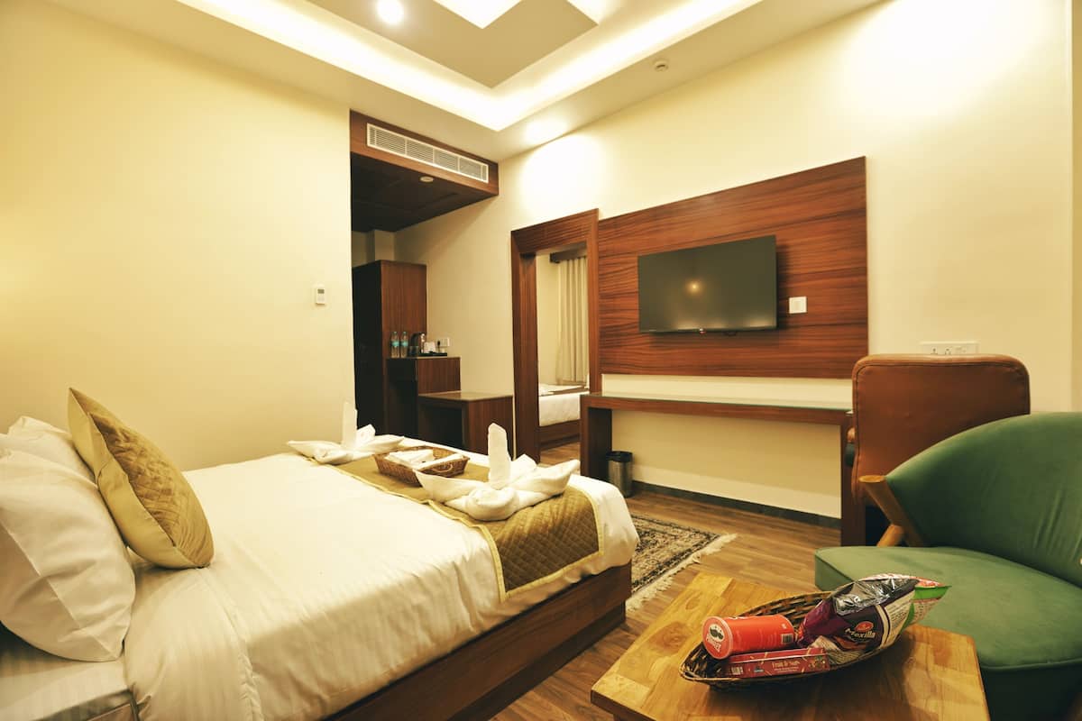 Quarto de hotel com uma cama, televisão fixada na parede, poltrona verde e mesa de cabeceira de madeira com cesta de lanches.