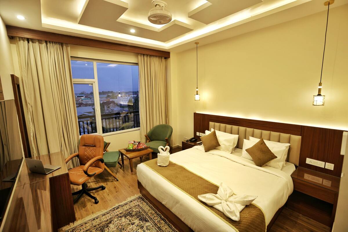 Quarto de hotel com uma cama grande, uma mesa com um laptop, uma cadeira, uma pequena mesa com lanches e vista para a cidade pela janela.