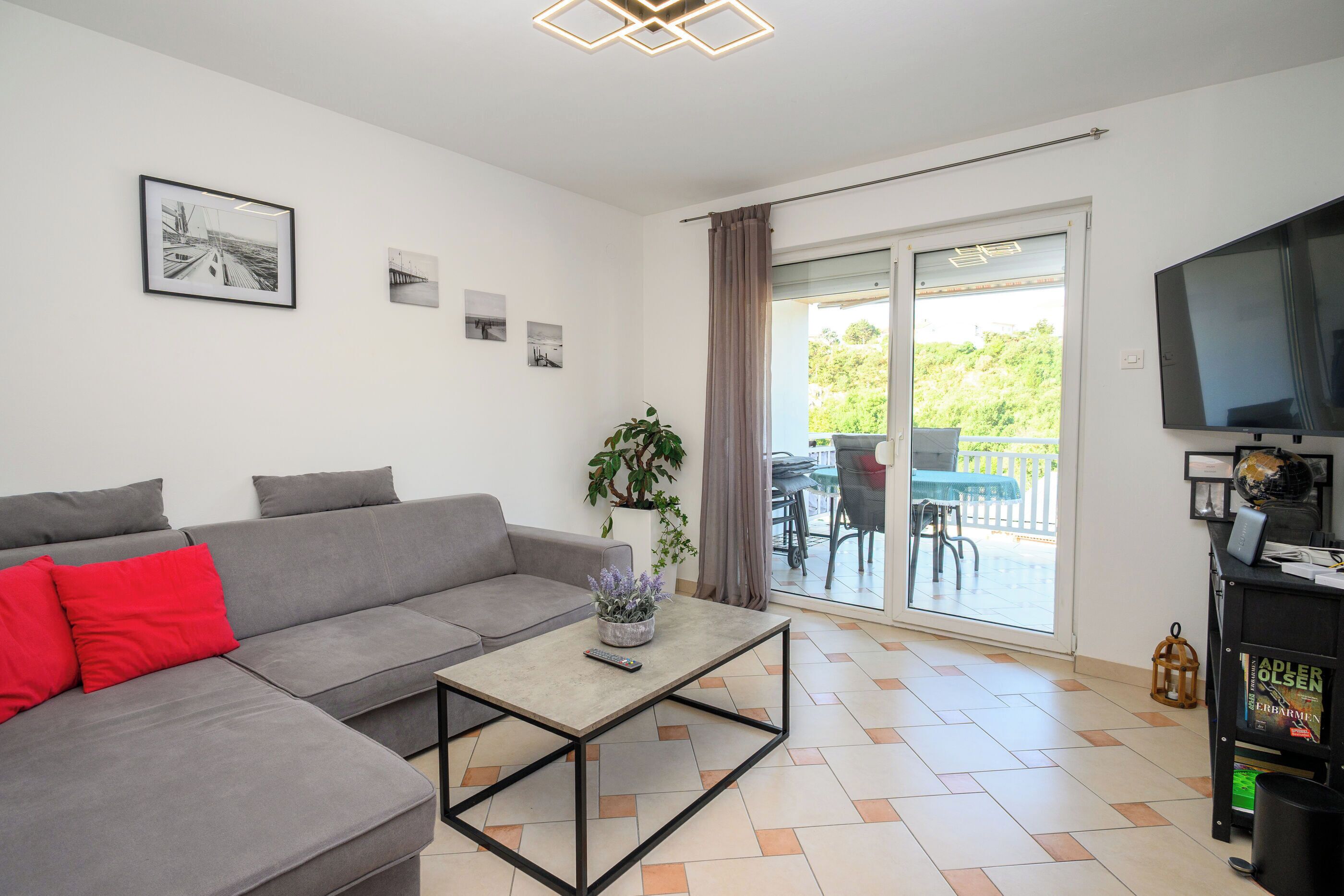 A modern living room with a grey sofa, a coffee table, a TV, and a balcony with outdoor furniture.