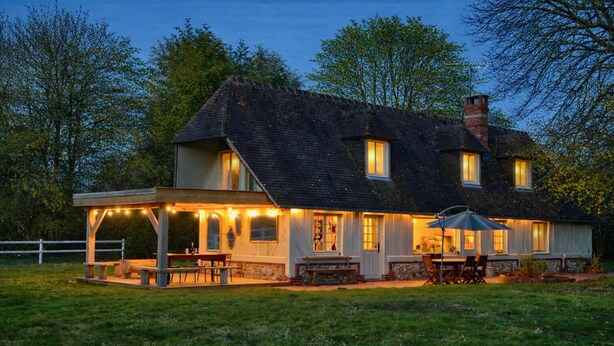 Ein zweistöckiges Haus mit überdachter Terrasse, Sitzgelegenheiten im Freien und einem gut beleuchteten Innenraum.