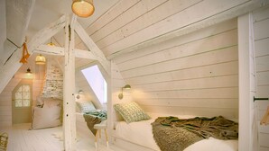 Ein Schlafzimmer im Loftsstil mit schrägem Dach, Holzbalken, einem Oberlicht, einem Bett mit Kissen, einem kleinen Hocker und einem Korb.