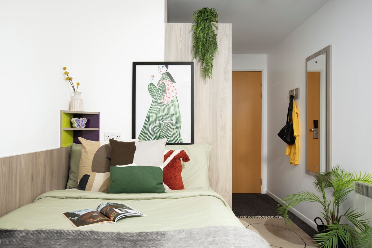 Ein modernes Schlafzimmer mit Bett, Kissen, einem gerahmten Kunstwerk, einer Zimmerpflanze, einem Bücherregal und einem Spiegel.