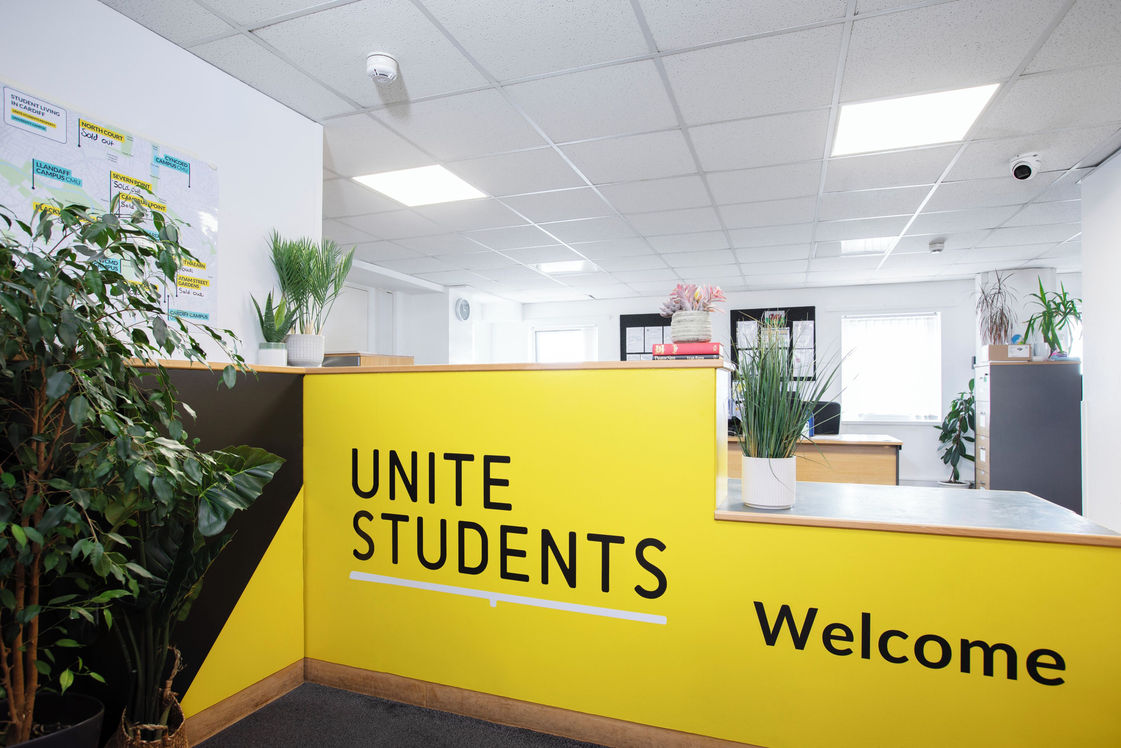 A reception area with a yellow counter, a sign reading 'UNITE STUDENTS Welcome', and potted plants.