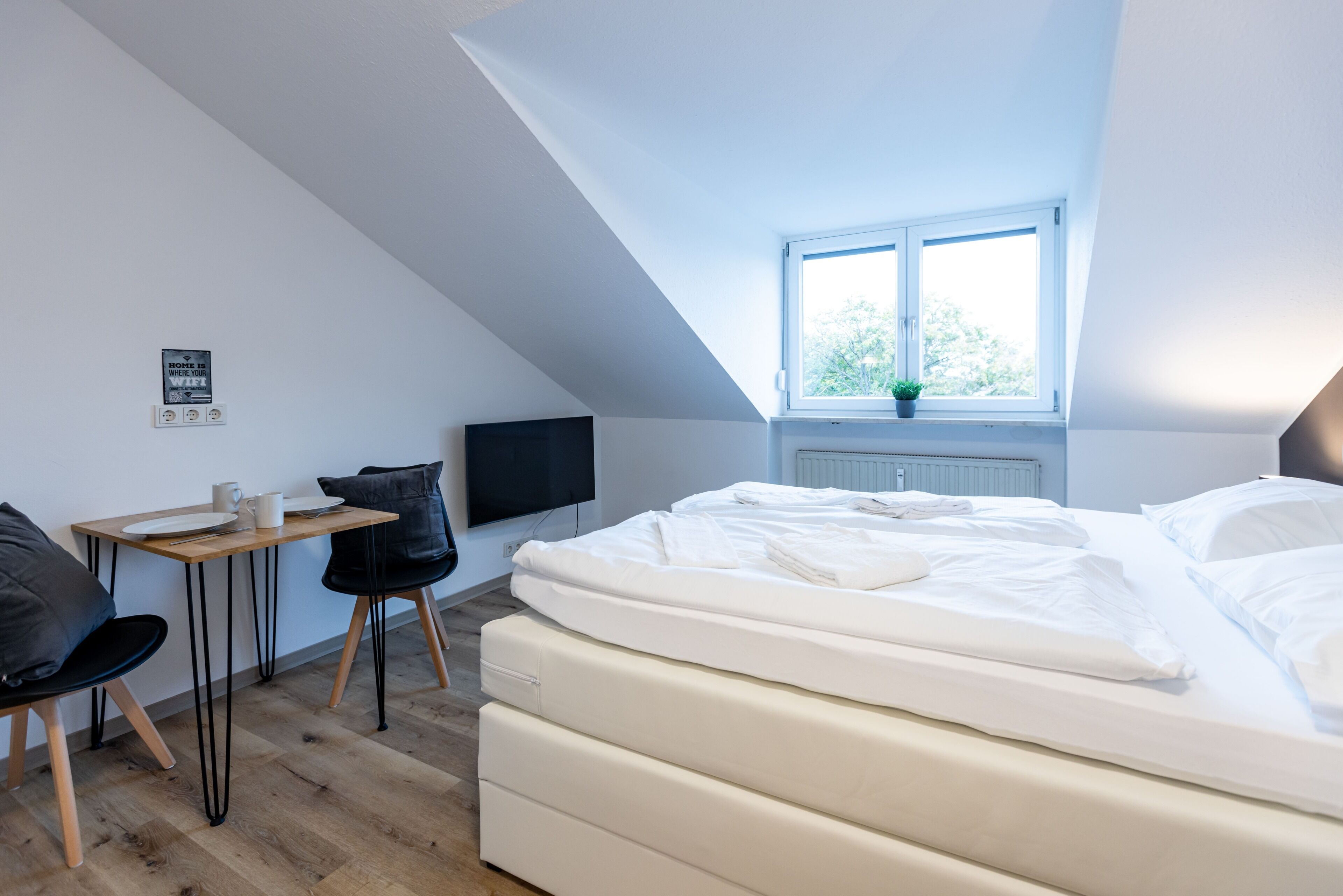 A bedroom with a bed, a desk, a chair, a television, and a window with a plant.