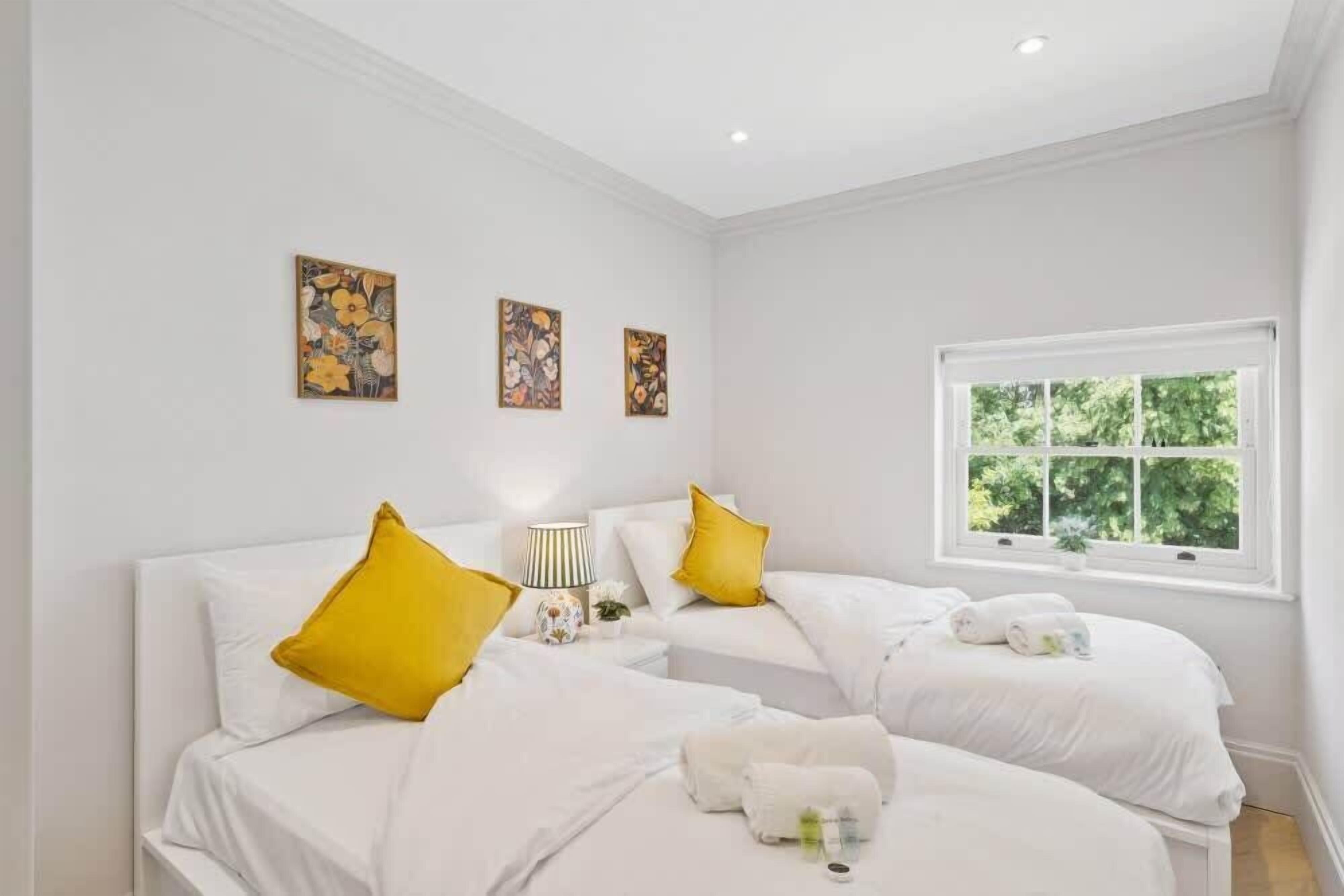 Habitación de hotel con dos camas, ropa de cama blanca, almohadas amarillas, una obra de arte en la pared, una ventana con vistas y una mesita de noche con una lámpara.