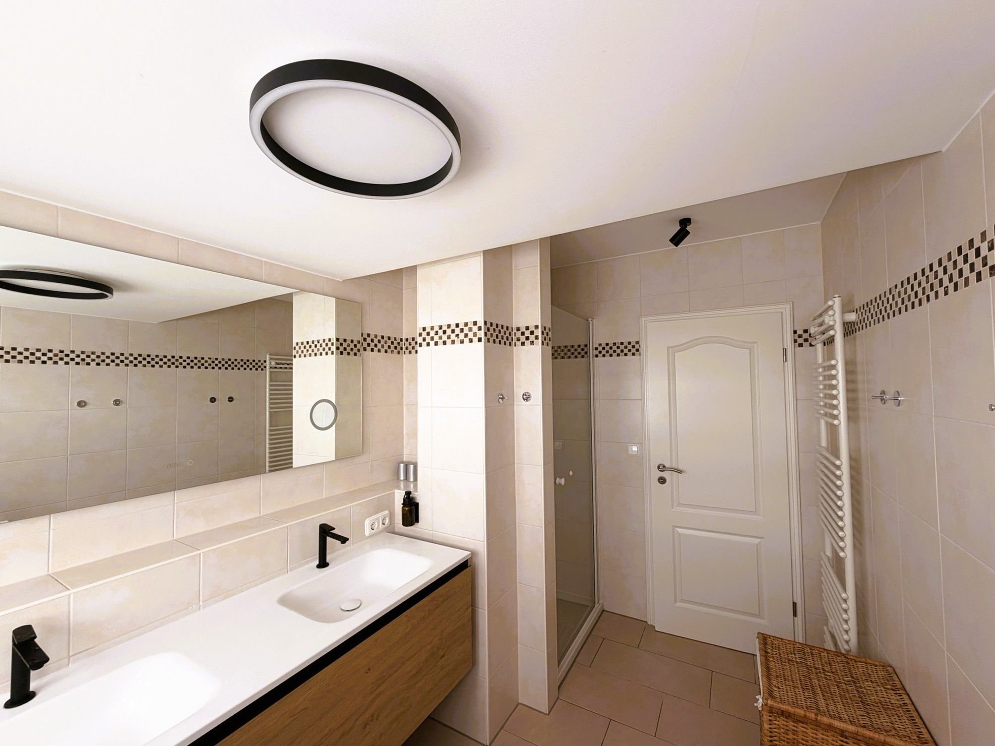 A modern bathroom with a double sink vanity, a large mirror, a shower area, and a door with a towel rack.