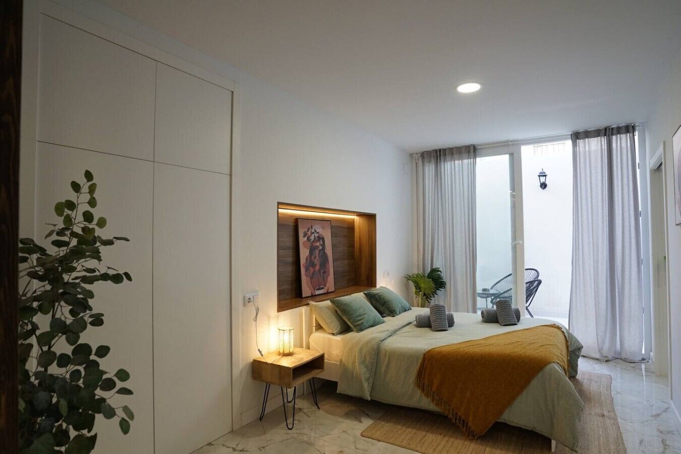 A bedroom with a bed, bedside table, lamp, and a plant.