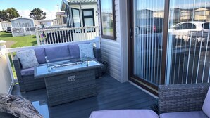 A deck with wicker furniture, a glass-top table, and a view of a parking area. - Sandy Beach Retreat (Prestatyn)
