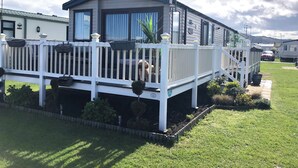 A mobile home with a white deck, a dog on the porch, and a car parked nearby. - Sandy Beach Retreat (Prestatyn)