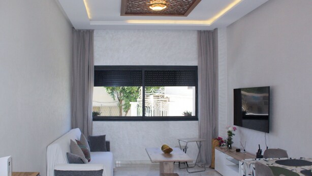 A modern room with a sofa, a dining table, a TV mounted on the wall, and a window with a view of the outside.