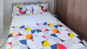 A small, single-bed room with a wooden wardrobe, a white air conditioning unit on the wall, and a colorful geometric-patterned bedspread.