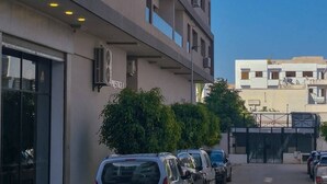 A multi-story building with a modern architectural design, featuring balconies, air conditioning units, and a parking area with several cars.