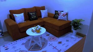 A living room with a brown sofa, a small white table with a vase, patterned rug, and a potted plant.