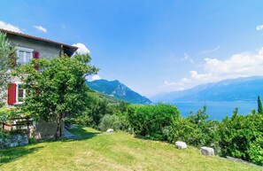 Ein Haus mit rotem Fensterlädchen, einer Steinmauer und Blick auf Berge und einen See.
