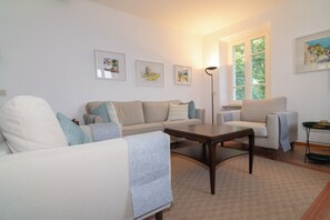 Ein Wohnzimmer mit einer Couch, Sesseln, einem Couchtisch und einer Stehlampe. An den Wänden hängen gerahmte Bilder.