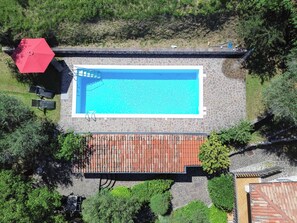Vista aérea de una piscina con un deck de piedra, rodeada de vegetación y un paraguas rojo.
