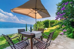 Ein Tisch mit Tischdecke, einer Flasche und Weingläsern, umgeben von Stühlen, mit Blick auf einen See und Berge im Hintergrund.