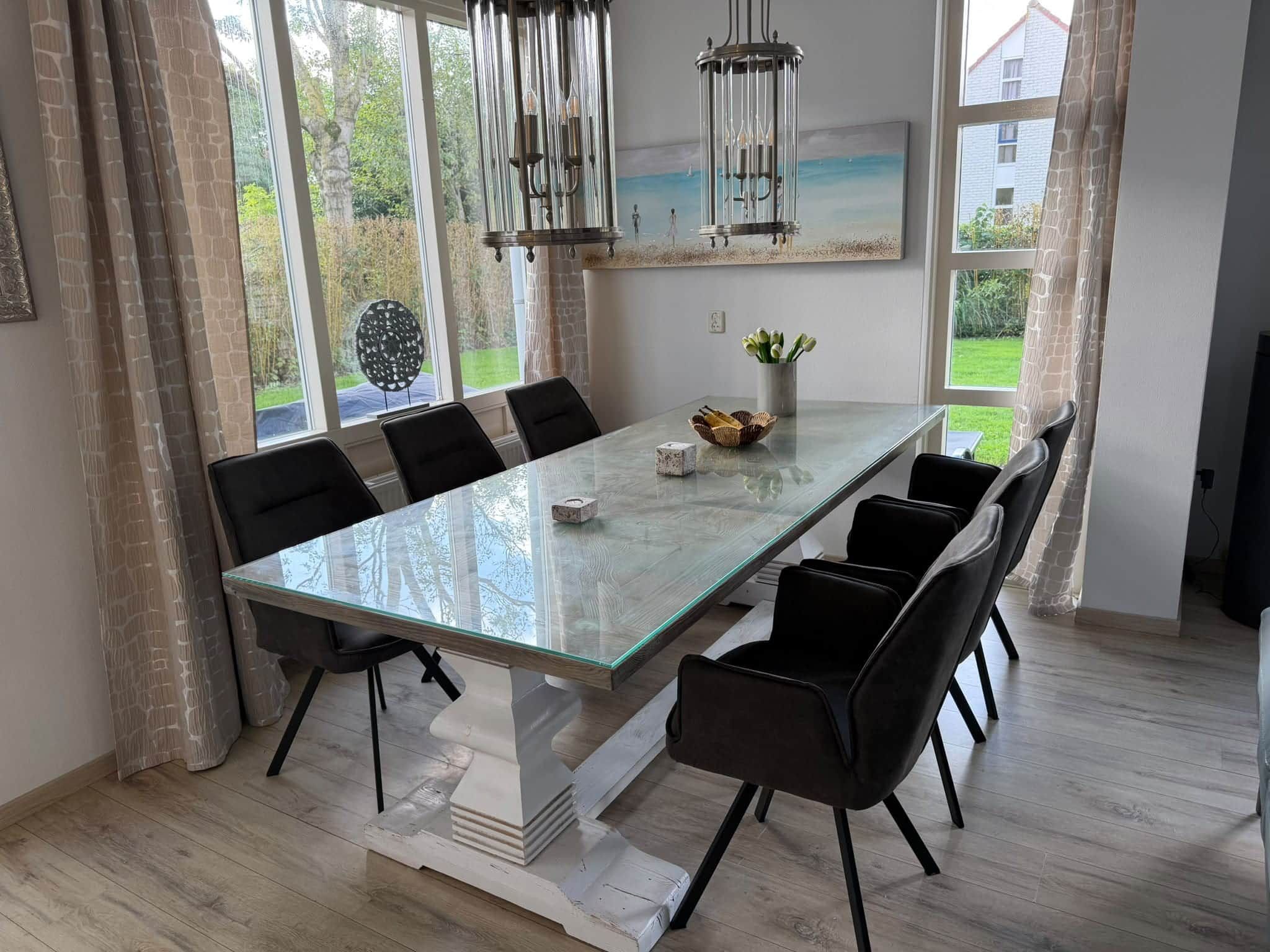 A dining room with a large glass table, black chairs, a chandelier, and a view of the outdoors.