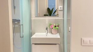 Casa de banho com lavatório branco, espelho, planta em vaso e chuveiro.