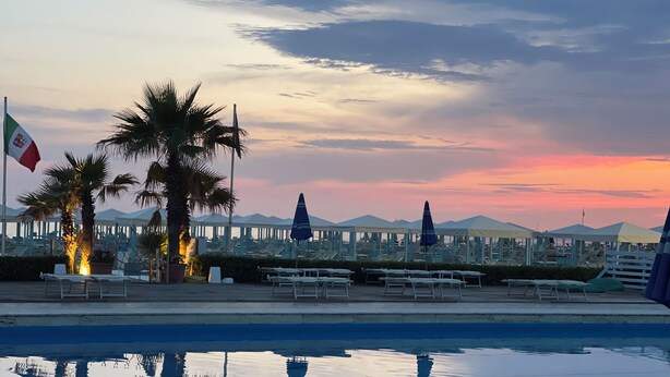 Piscina à beira-mar com espreguiçadeiras e guarda-sóis, palmeiras e vista para o pôr do sol.
