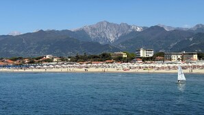 Uma vila costeira com uma praia, um veleiro e montanhas ao fundo.