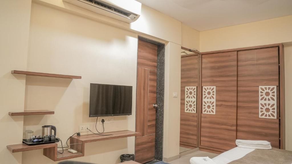 Una habitación de hotel con televisor de pantalla plana, un armario de madera con paneles decorativos, una pequeña estantería con hervidor y una mesita de noche con toalla.