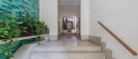 Una escalera con barandilla de madera, pared con azulejos y vegetación, y una vista despejada hacia un cuarto con puerta de vidrio.