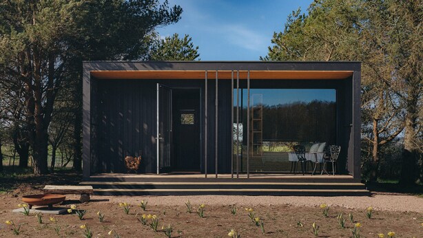 A modern building with a large glass window, a wooden deck, and a garden with yellow flowers. - Curlew (St. Andrews)