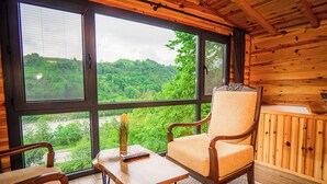 Interior de una cabaña de madera con un ventanal grande, un sillón con cojines, una mesita con un florero y un control remoto, y un armario de madera.
